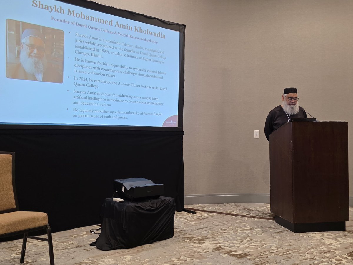 Speaker in traditional Islamic attire stands at podium during presentation about Shaykh Mohammed Amin Khatiwada projected on screen