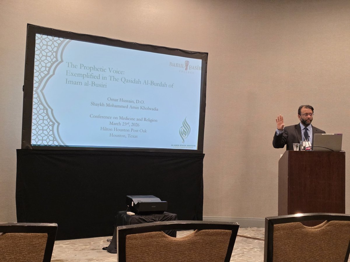 Man in suit speaking at podium with presentation displayed on large screen behind him in conference room