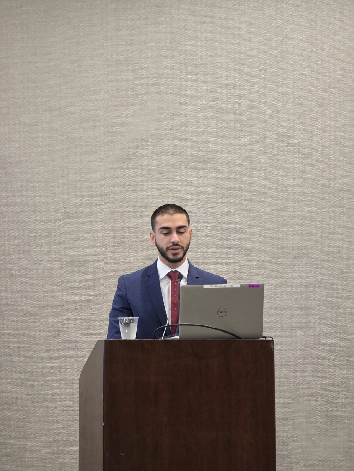 Man in blue blazer and red tie speaking at podium with laptop and water glass